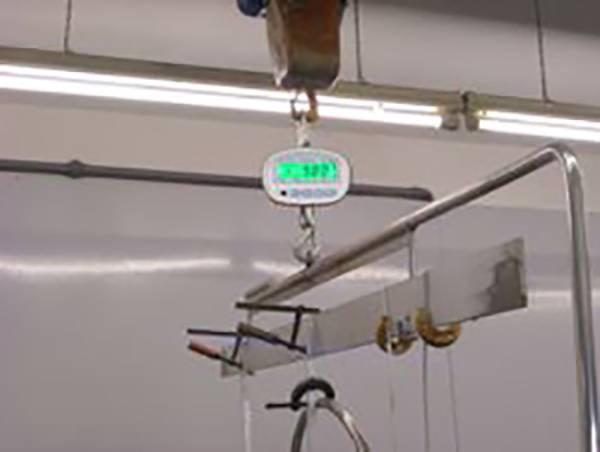 Een industrieel weegtoestel hangt aan een haak en geeft het gewicht aan van een object dat bevestigd is aan een metalen staaf.