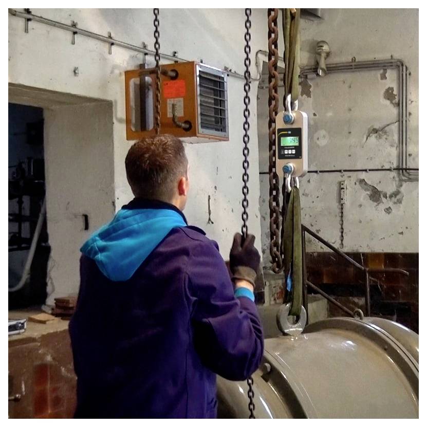Arbeider in een werkplaats bedient een kettingblok om een grote metalen cilinder die aan kettingen hangt omhoog te tillen.