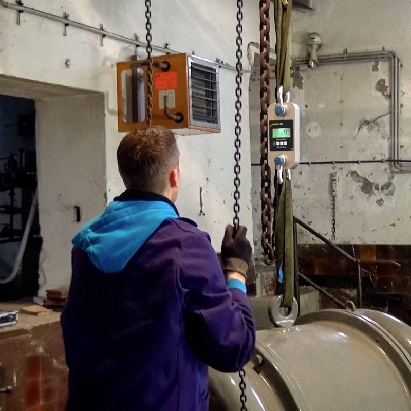 Een man in werkkleding bedient een industriële hijsinrichting. Een weergaveapparaat aan de haak toont een gewicht aan.