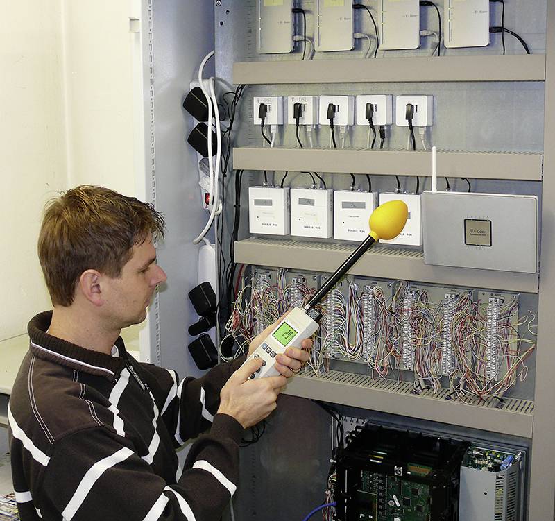 Een man in een gestreepte trui meet met een geluidsniveaumeter de geluiden bij een open schakelkast vol kabels en apparaten.