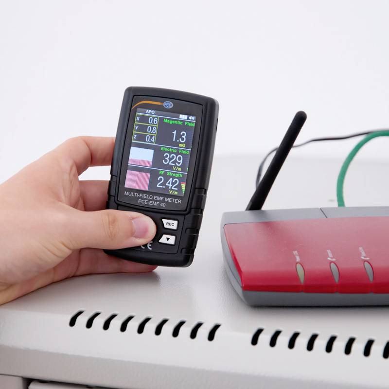 Een persoon houdt een EMF-meetapparaat naast een router om elektromagnetische velden te meten.