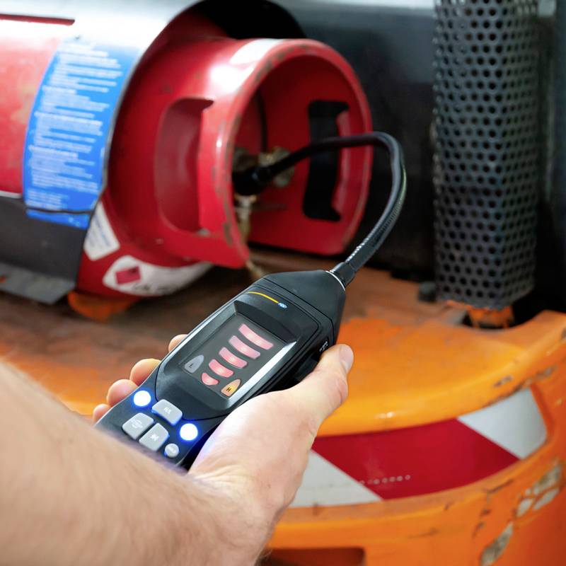 Een persoon houdt een gasmetingsapparaat voor een rode gasfles om het gasniveau of lekken te controleren. Op de achtergrond een industriële omgeving.
