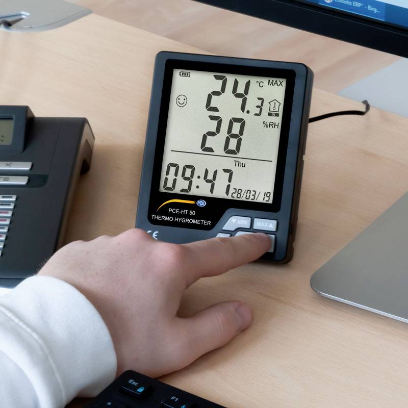 Een persoon bedient een digitaal thermo-hygrometer dat 24,1°C en 28% luchtvochtigheid aangeeft. Een telefoon en een monitor zijn op de achtergrond zichtbaar.