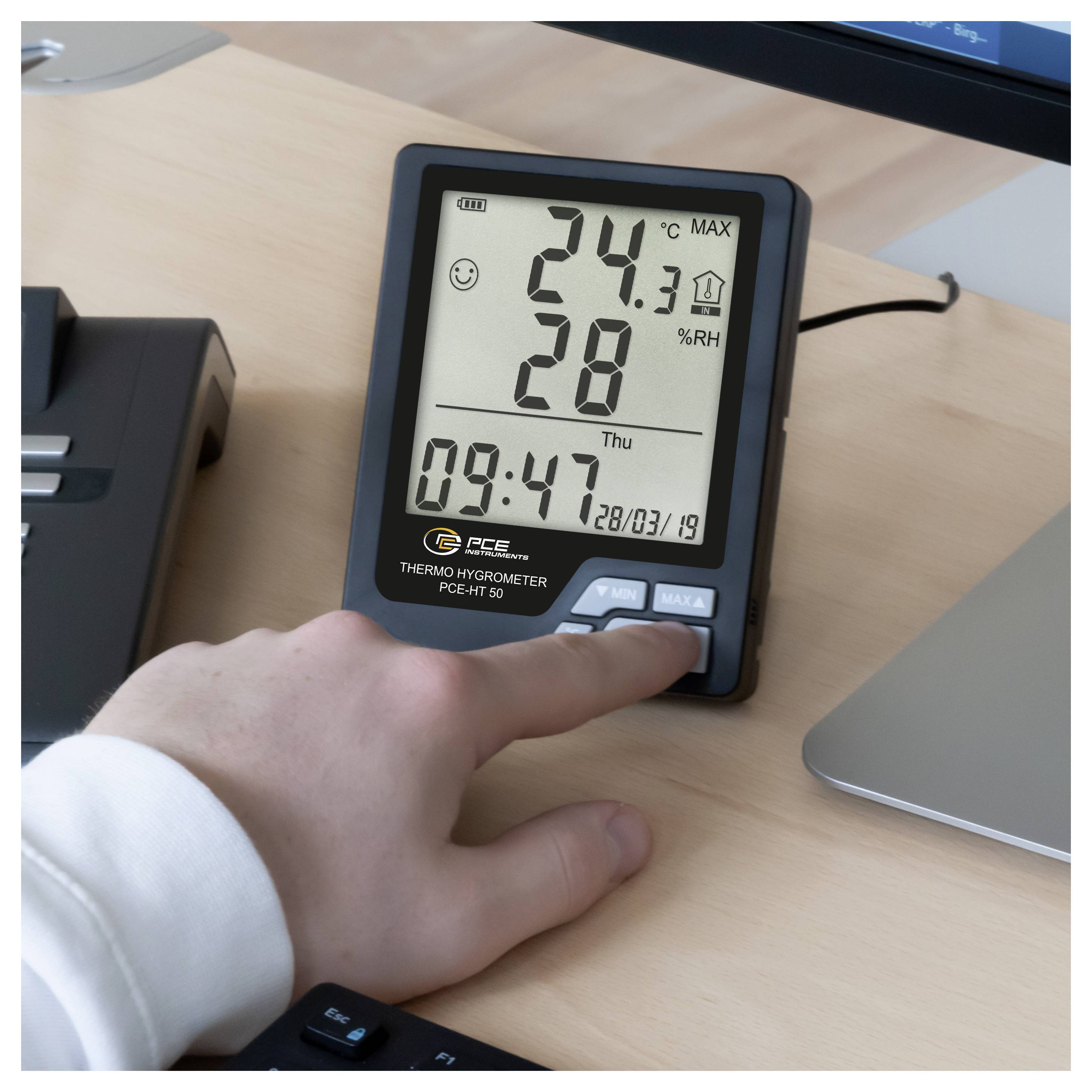 Hand drukt op de 'MAX/MIN'-knop van een digitale thermo-hygrometer die 24,3°C en 28% vochtigheid op een bureau weergeeft.