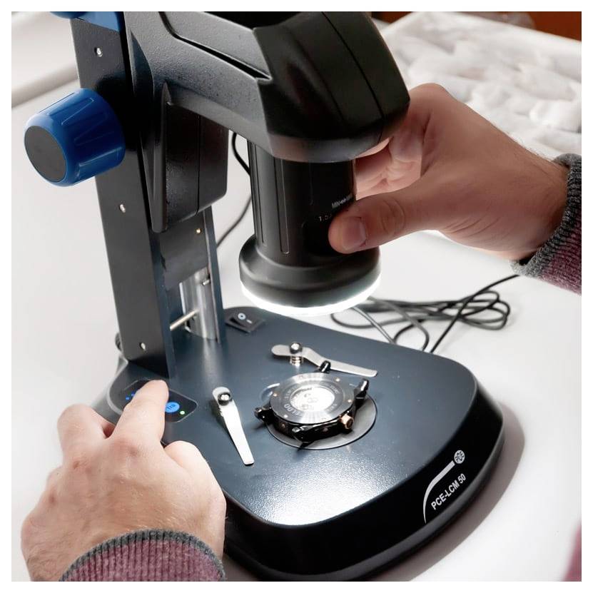 Handen die een digitale microscoop op een labbank aanpassen, met de verlichte lens gericht op het podium en monsterbereik.