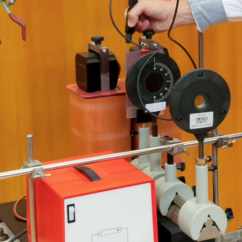 Een hand bedient een precisiemeetinstrument in een laboratorium. De omgeving bevat technische apparatuur en meetschalen.