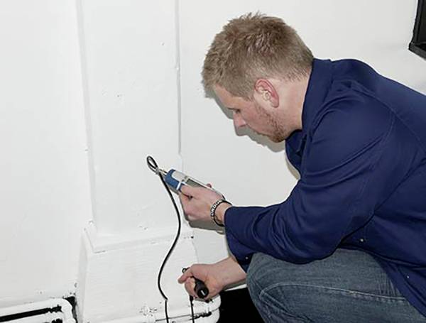 Een man in werkkleding gebruikt een meetapparaat tegen een witte muur. Hij meet waarschijnlijk vocht of andere parameters.