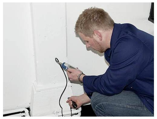 Een man hurkt bij een muur en gebruikt een handheld gereedschap om een elektrisch stopcontact en kabel nabij de vloer te inspecteren of testen.