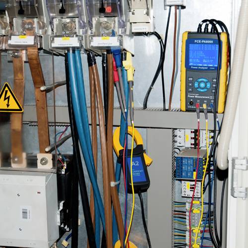 Elektrische schakelkast met verschillende kabels en een aangesloten meetapparaat voor spanning- en stroomcontrole.