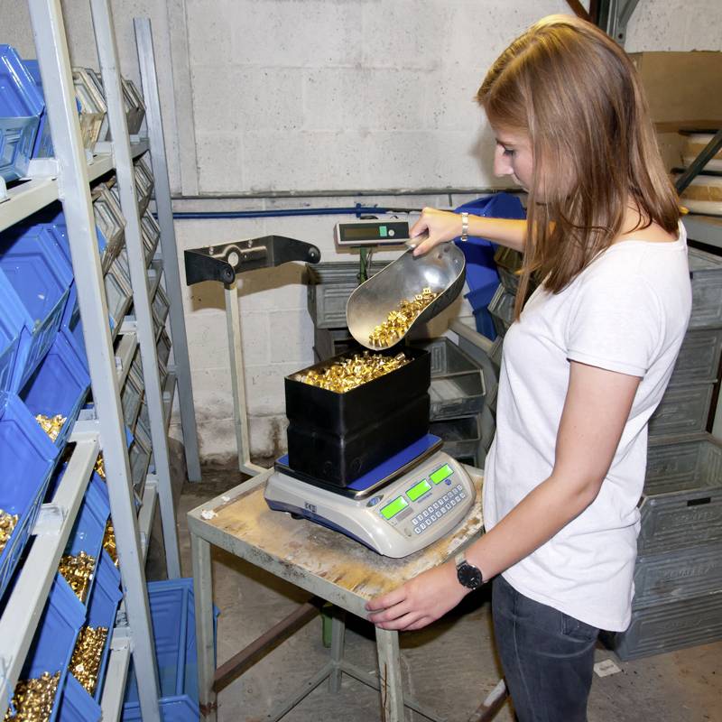 Een vrouw weegt een grote hoeveelheid gouden patronenhulzen op een digitale weegschaal in een industriële omgeving.
