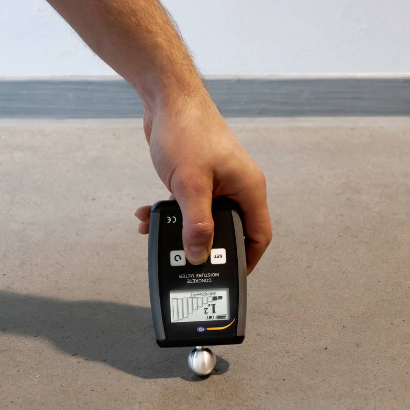 Een hand houdt een vochtmeter vast op een betonvloer. Het display toont een balkwaarde.