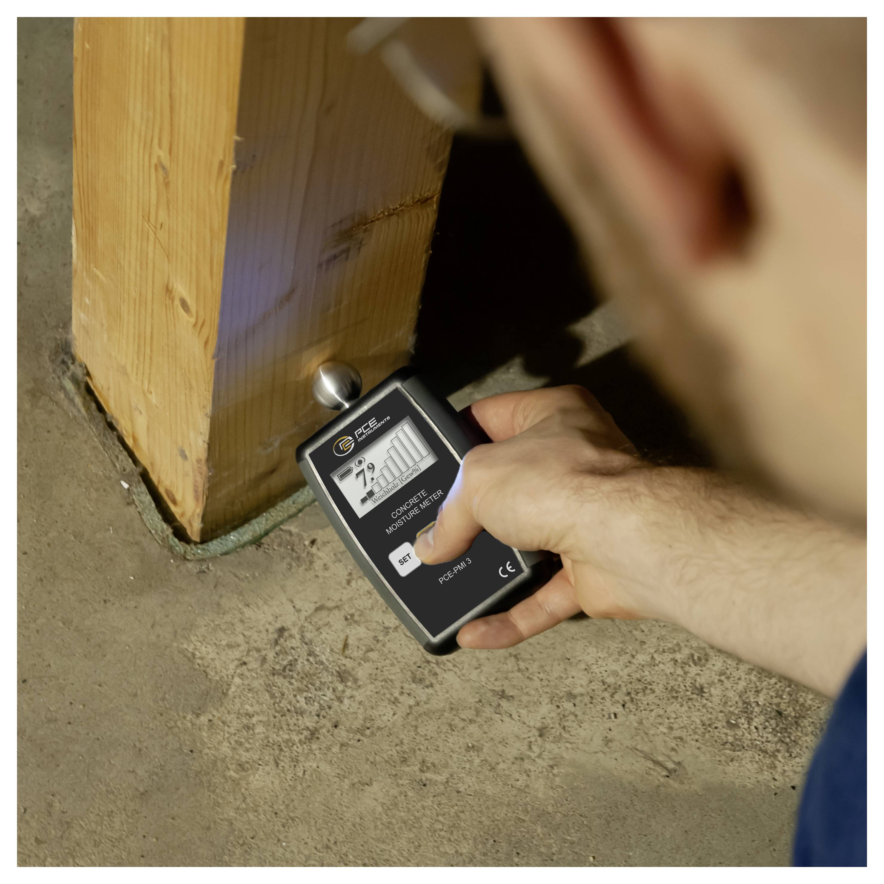 Persoon die een handheld vochtmeter gebruikt om de basis van een houten paal te controleren waar deze op de betonnen vloer aansluit.