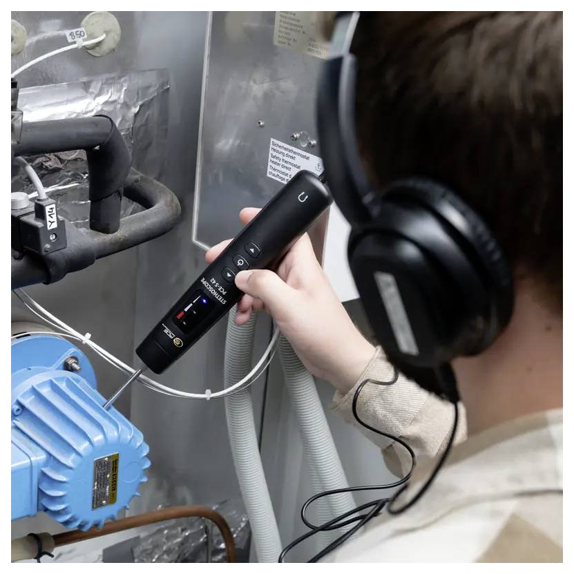 Een persoon die koptelefoons draagt, gebruikt een handheld apparaat om machines met blauwe en zwarte componenten te inspecteren, wat op een technische operatie wijst.