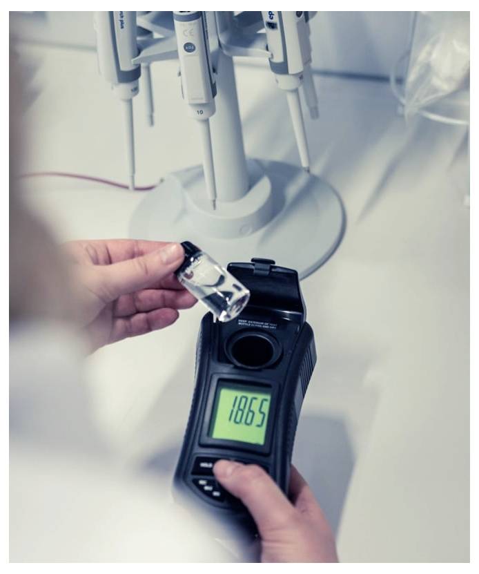 Een persoon meet een klein flaconnetje met een handheld digitale meter in de buurt van laboratoriumpipmenten, met een uitlezing van 1865.
