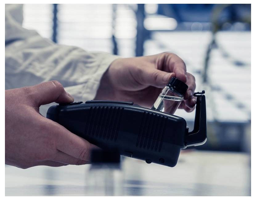 Handen voegen een klein flesje in een zwart handheld apparaat in, waarschijnlijk een monsterschanner of laboratoriumapparaat, in een wazig laboratoriummilieu.