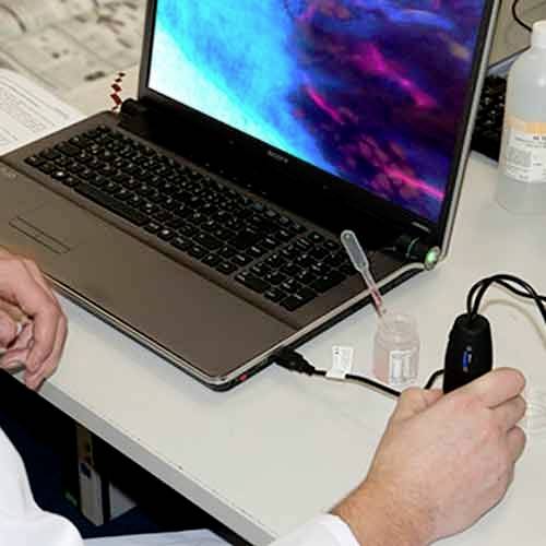 Een persoon voert een experiment uit op een laptop, met een aangesloten microscoop en een kleine flesje vloeistof.