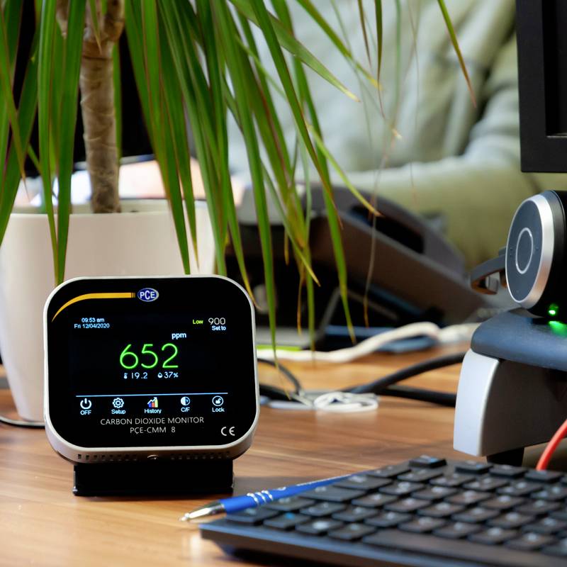 Een tafel met een CO2-monitor die 652 ppm aangeeft, geplaatst naast een pc-monitor. Een plant staat op de achtergrond.