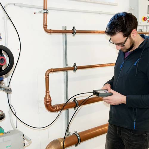 Een man staat in een technische ruimte en controleert een apparaat. Achter hem zijn buizen en een drukmeter zichtbaar.