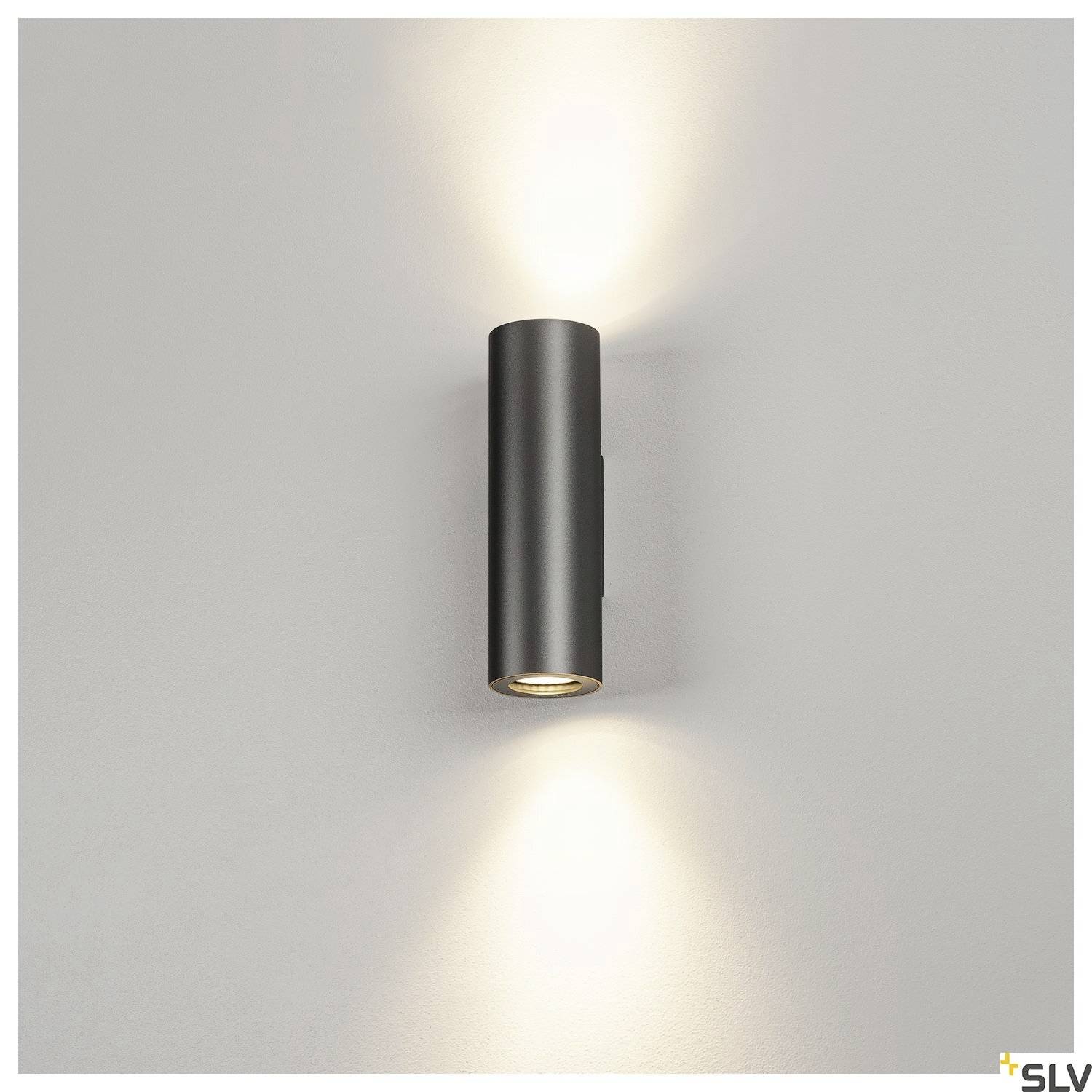 Wandlamp in cilindrische vorm aan een grijze muur, straalt licht naar boven en beneden, creëert warm licht en zachte schaduwen.