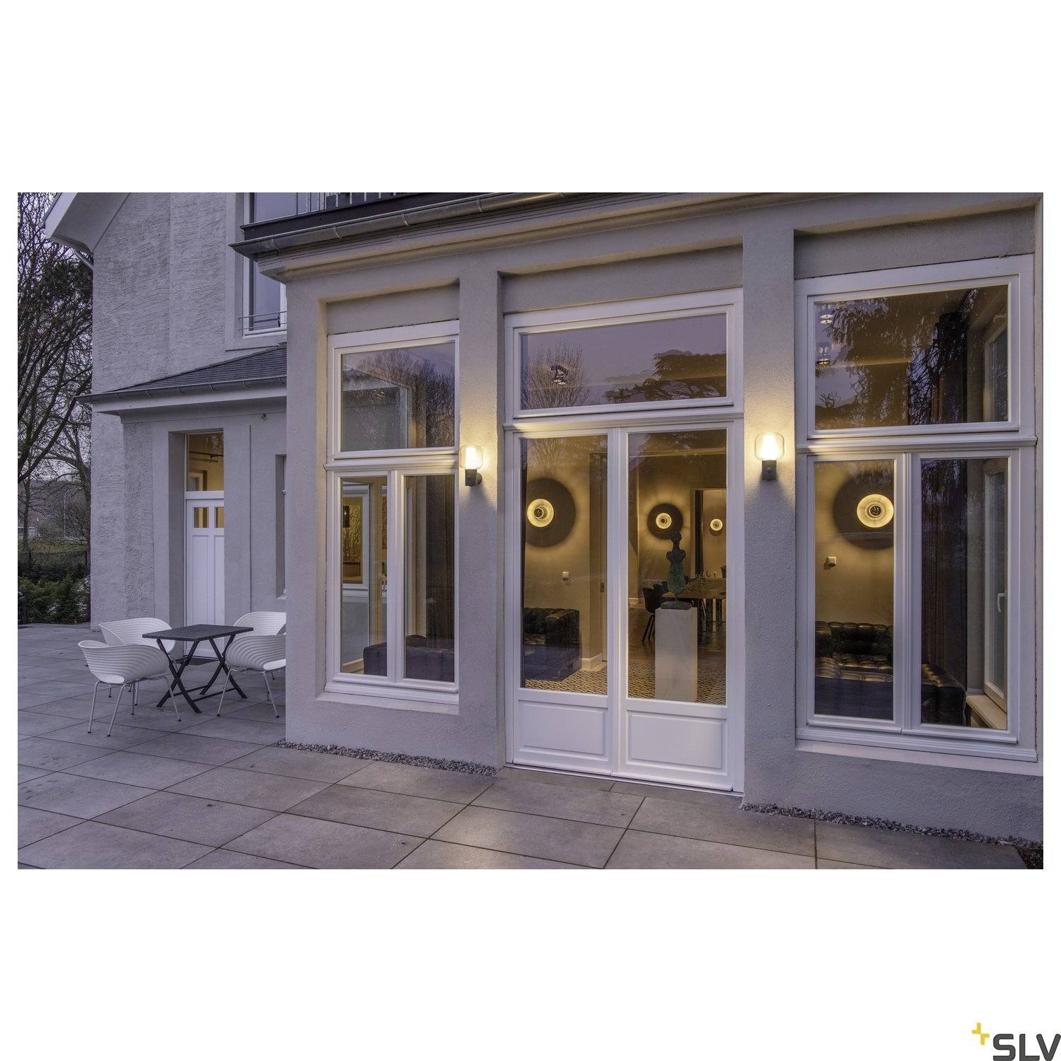 Huis met verlichte ingangspartij. Twee ronde buitenlampen naast de deur en ramen. Terras met tafel en twee stoelen.