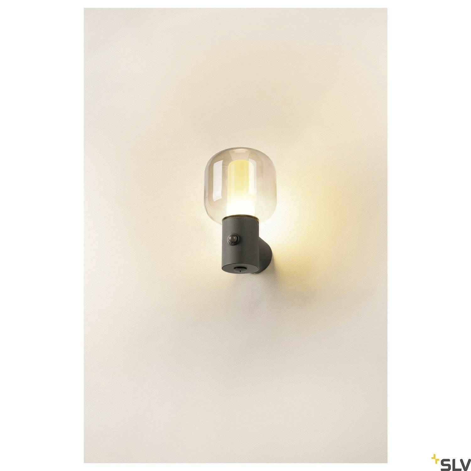 Eenvoudige wandlamp met een zwarte voet en een heldere glazen kap, die warm licht op een witte muur werpt.