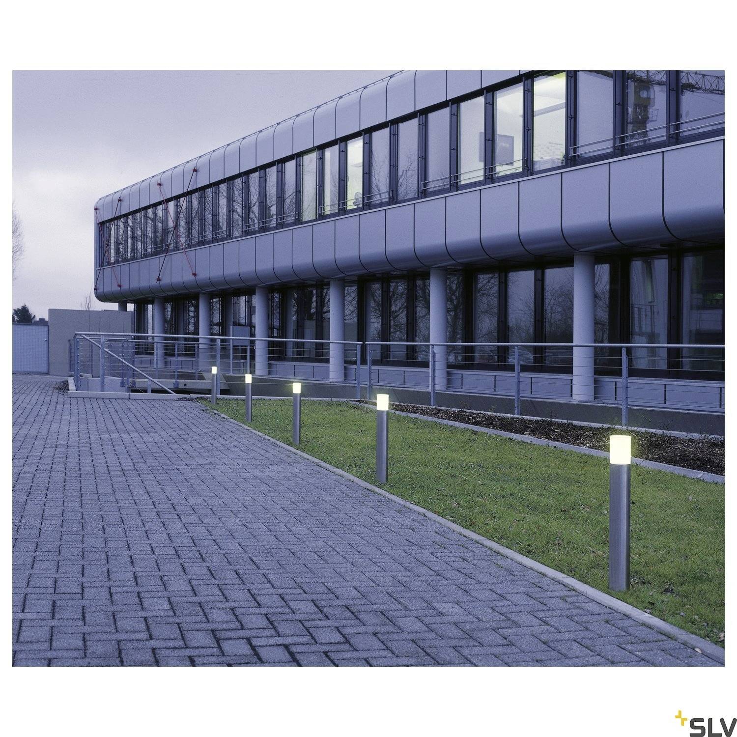 Een modern kantoorgebouw met een glazen gevel bij het invallen van de avond. De voorgrond toont een geplaveid voetpad met verlichte wegverlichting.