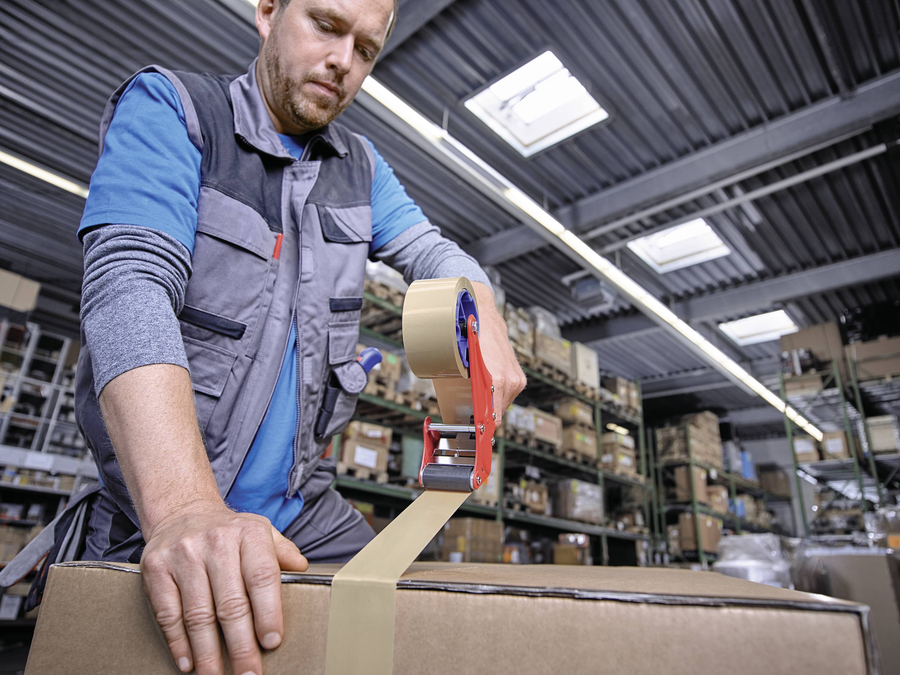 Een magazijnmedewerker sluit een grote doos af met plakband in een magazijn, omringd door stellingen vol dozen en goederen.