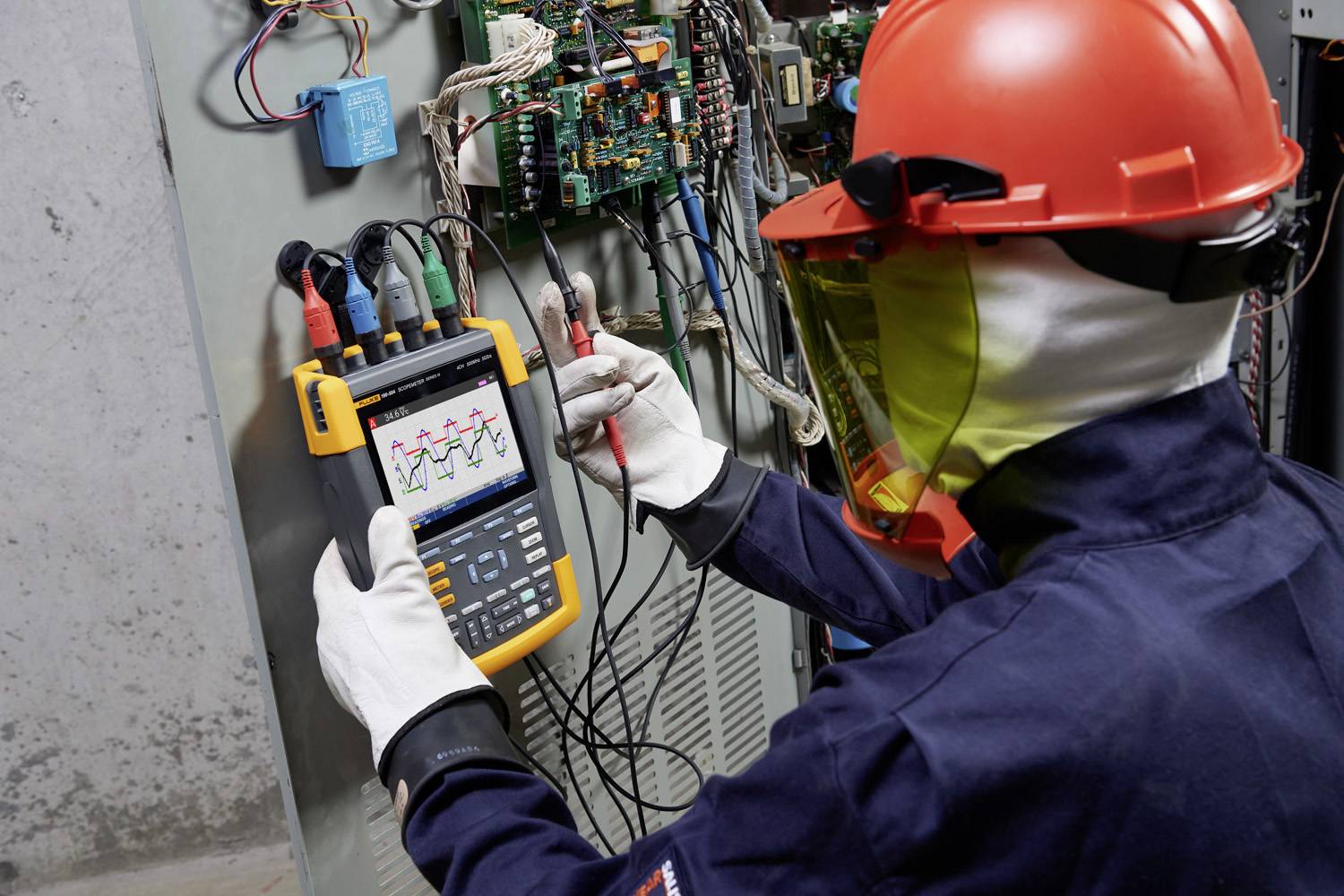 Een technicus met veiligheidshelm en -masker controleert elektrische schakelingen met een oscilloscoop in een schakelkast.
