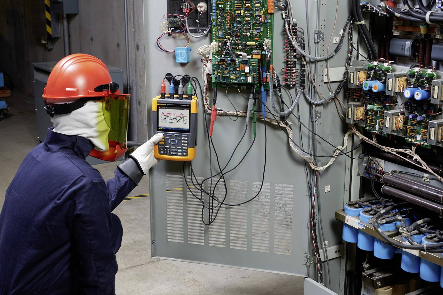 Een persoon in beschermende uitrusting meet elektrische signalen aan een open schakelkast met een draagbaar oscilloscoop in een industriële omgeving.