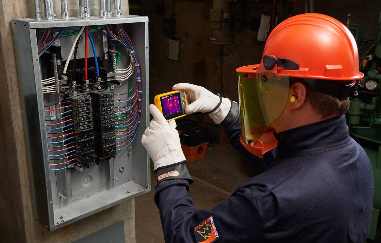 Een technicus in beschermende uitrusting gebruikt een warmtebeeldcamera voor inspectie van een open zekeringkast om elektrische problemen te detecteren.