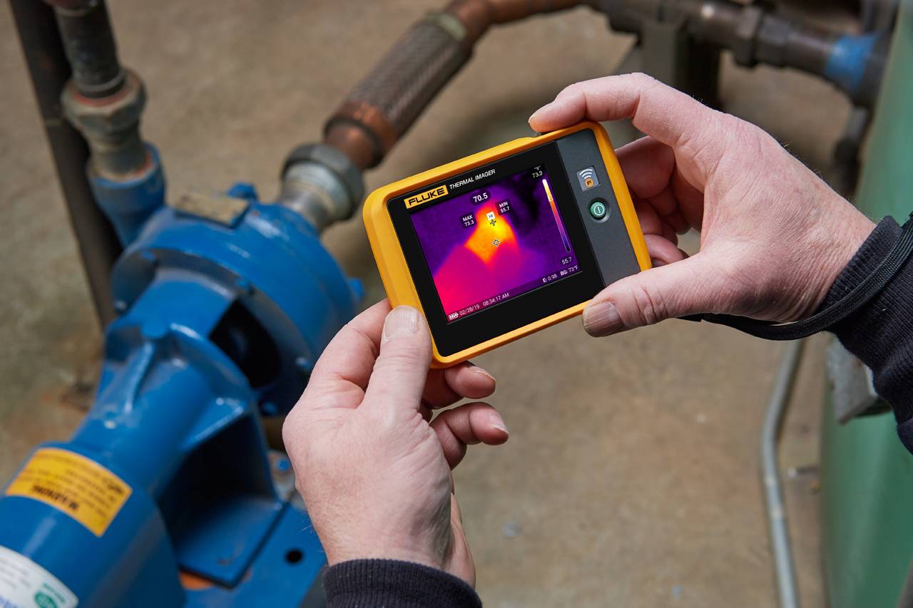 Een persoon houdt een warmtebeeldcamera en meet de temperatuur van een blauwe machinemotor. Het display toont een thermisch beeld.
