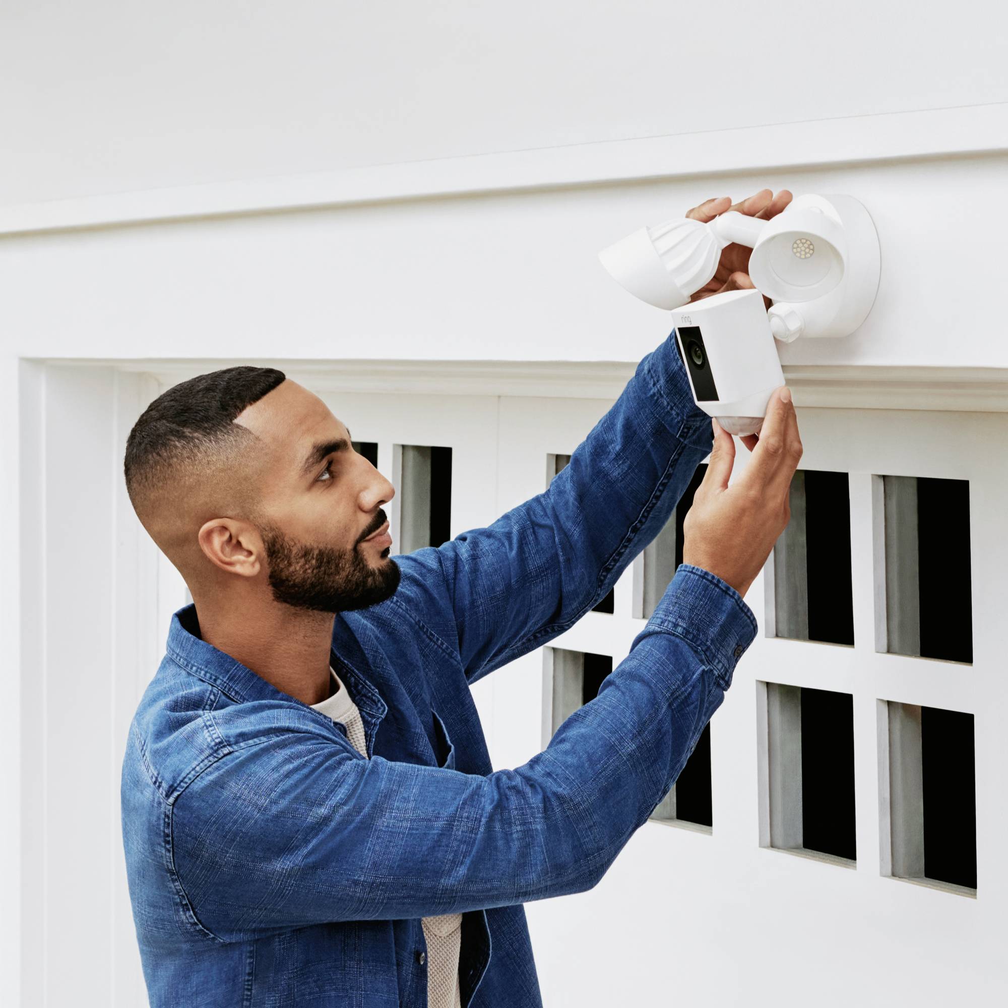 Een man in een spijkerblouse installeert een witte buitencamera aan de muur van een huis, kijkend omhoog, met de focus op beveiligingsmaatregelen.