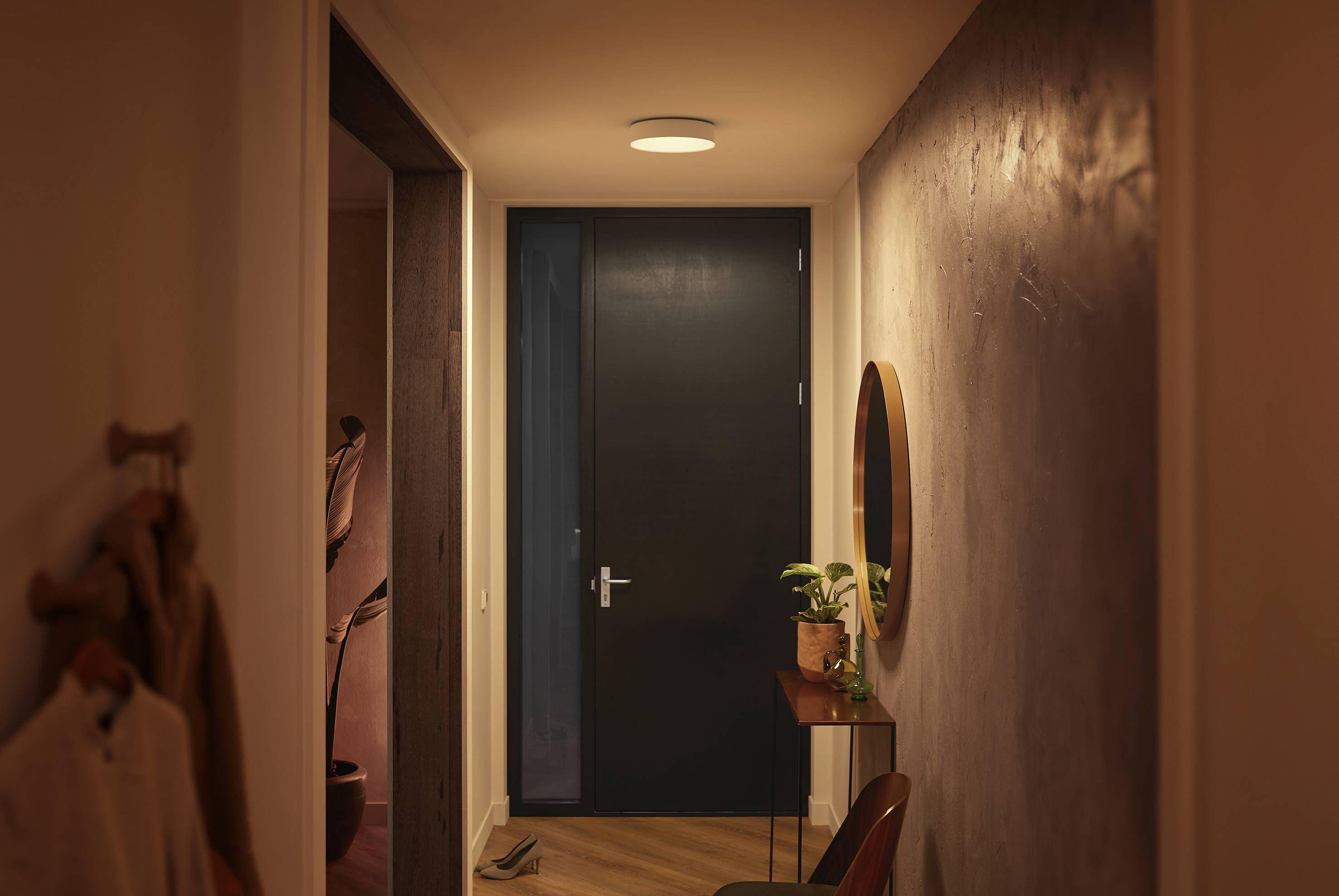 Hal met warm licht, waaraan een gesloten donkere deur zich aan het einde bevindt. Rechts een ronde spiegel boven een kleine tafel met een plant.