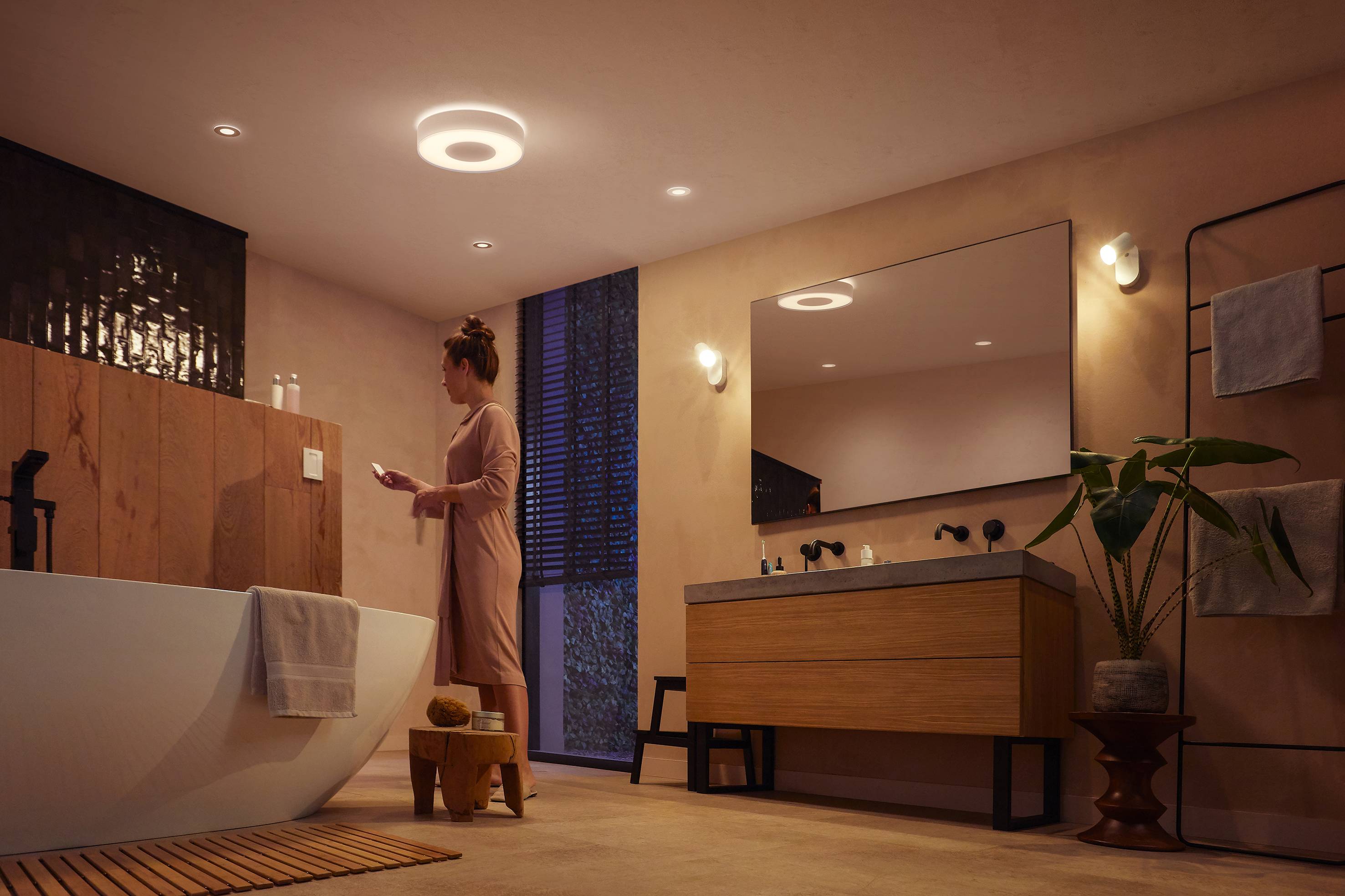 Een persoon staat in een modern badkamer met een badkuip, wastafel en planten. De verlichting creëert een warme sfeer.