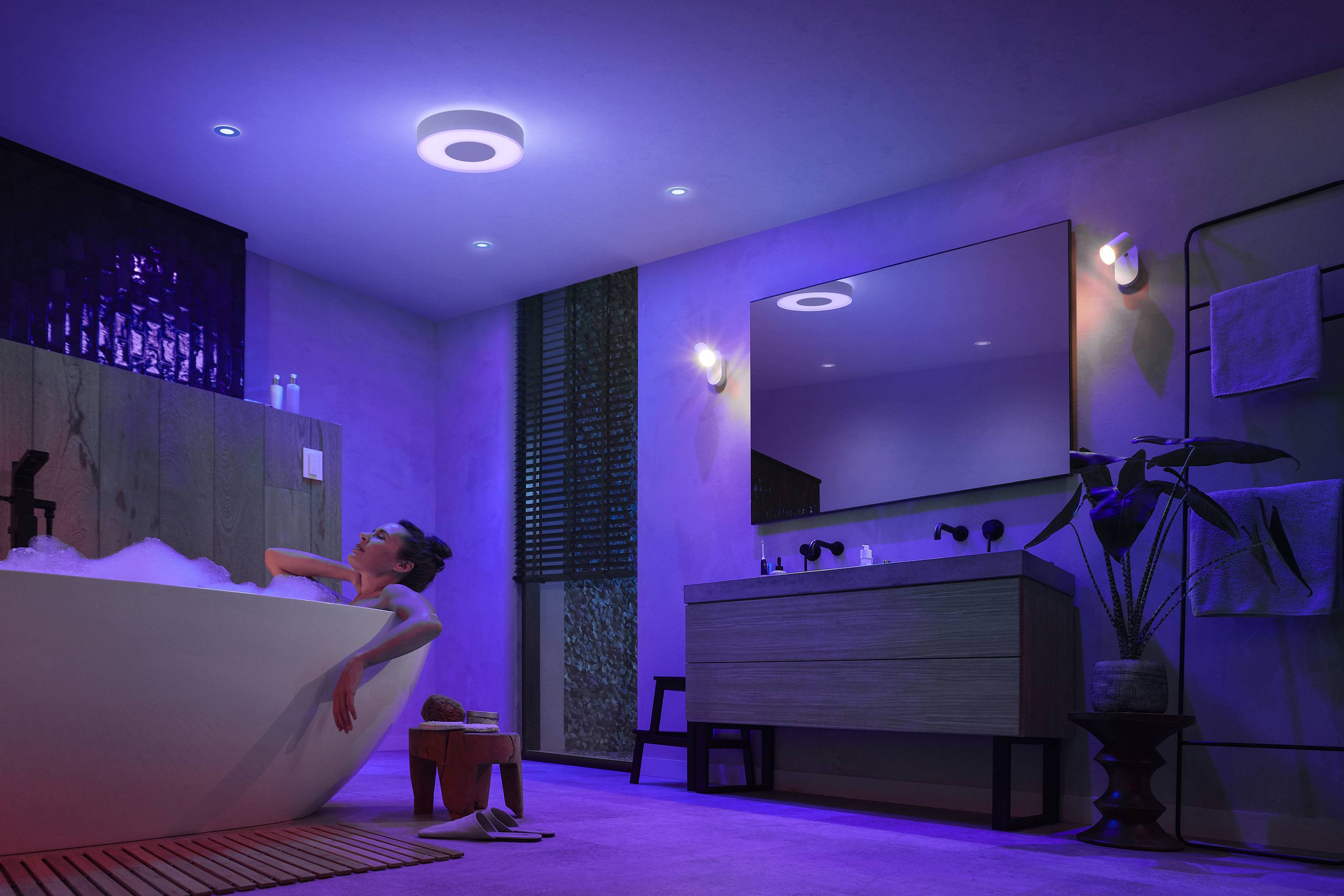 Een persoon ontspant in een badkuip in een sfeervol verlichte badkamer. De verlichting schakelt tussen blauw en paars.