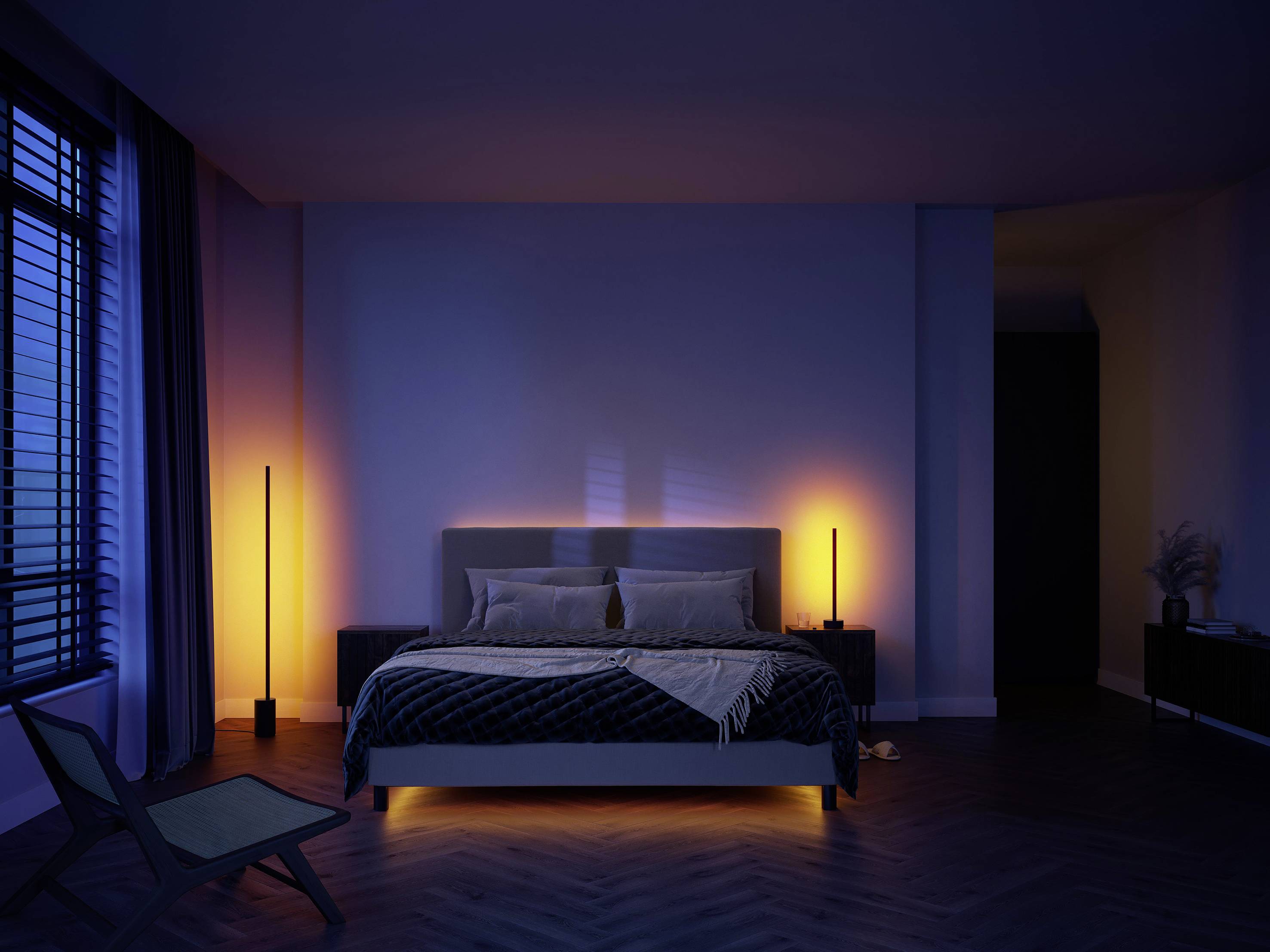 Een slaapkamer met een groot bed, verlicht door warm licht. Links staan twee hoge lampen. Rechts een stoel voor een raam.