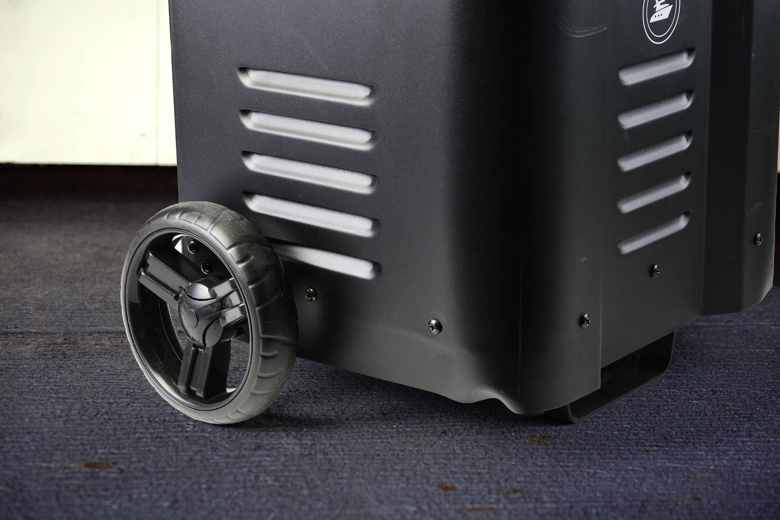 Zwarte transportbox op blauwe tapijtvloer met een groot wiel, geschikt voor gemakkelijk transport van apparatuur of bagage.