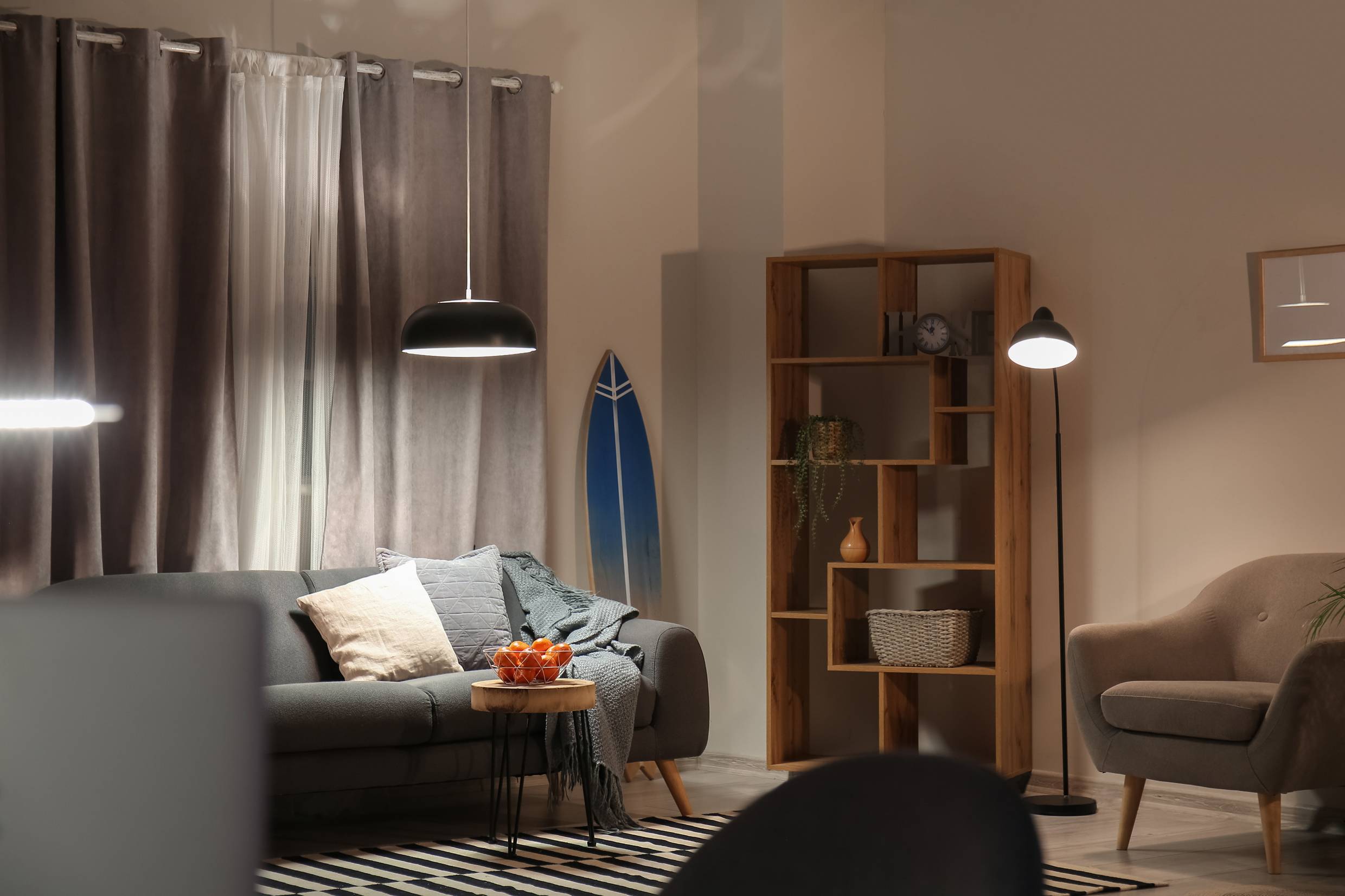 Gezellige woonkamer met een grijze bank, sierkussens, bijzettafel met fruit, surfplank aan de muur, staande lamp en boekenplank.