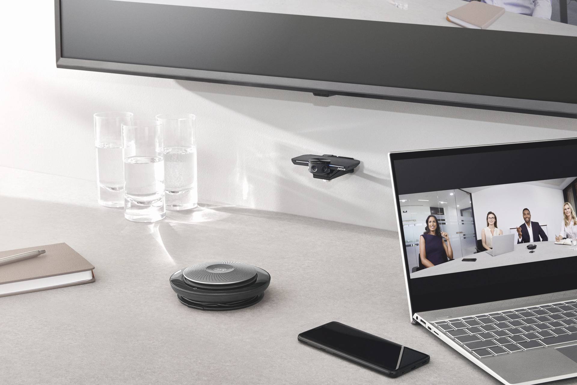 Vergaderzaal met laptop die een videoconferentie met vijf personen weergeeft. Op de voorgrond: smartphone, notitieboek, microfoon.