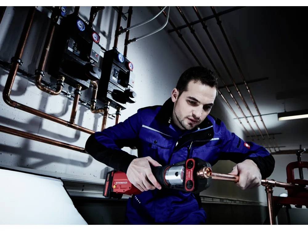 Een persoon in een blauwe uniform gebruikt een rode elektrische gereedschap om te werken aan koperen leidingen in een industriële omgeving, omringd door meters en pijpen.