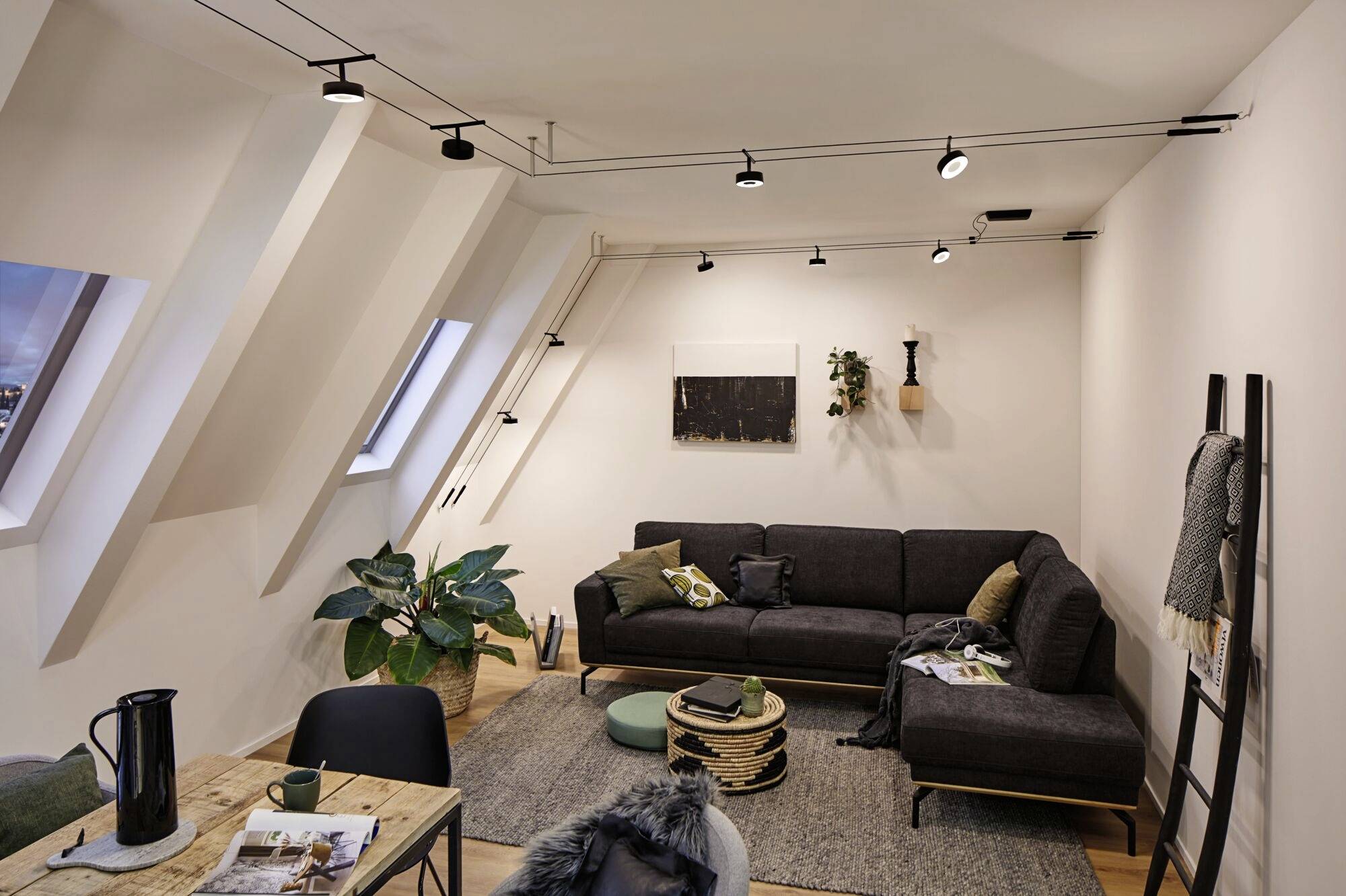 Woonkamer op zolder met schuine wanden, grijze bank, planten en houten tafel. Modern design met dakramen en verlichting.