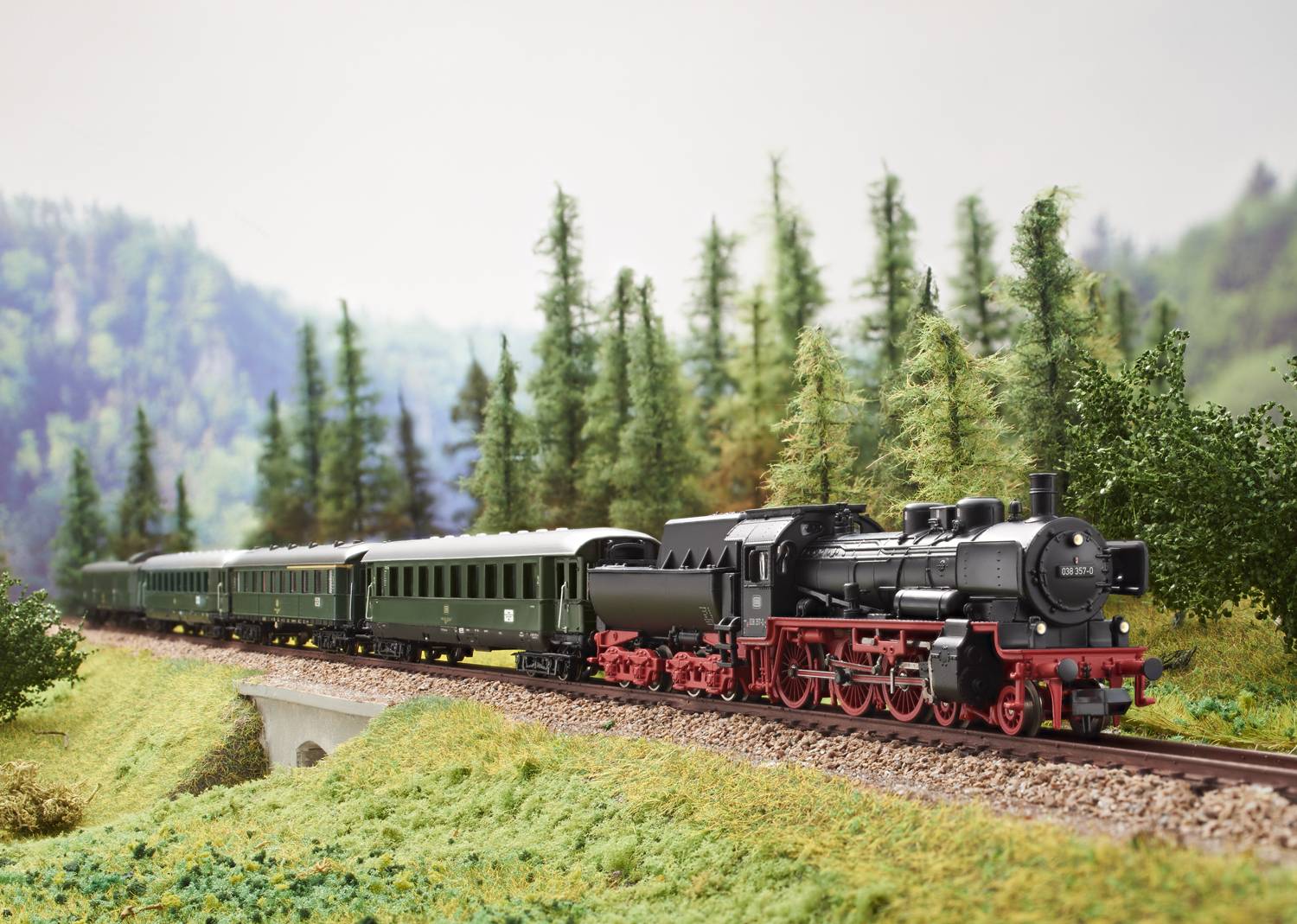 Een modeltreintje rijdt door een landschap met bomen en heuvels, bestaande uit een stoomlocomotief en groene wagons.