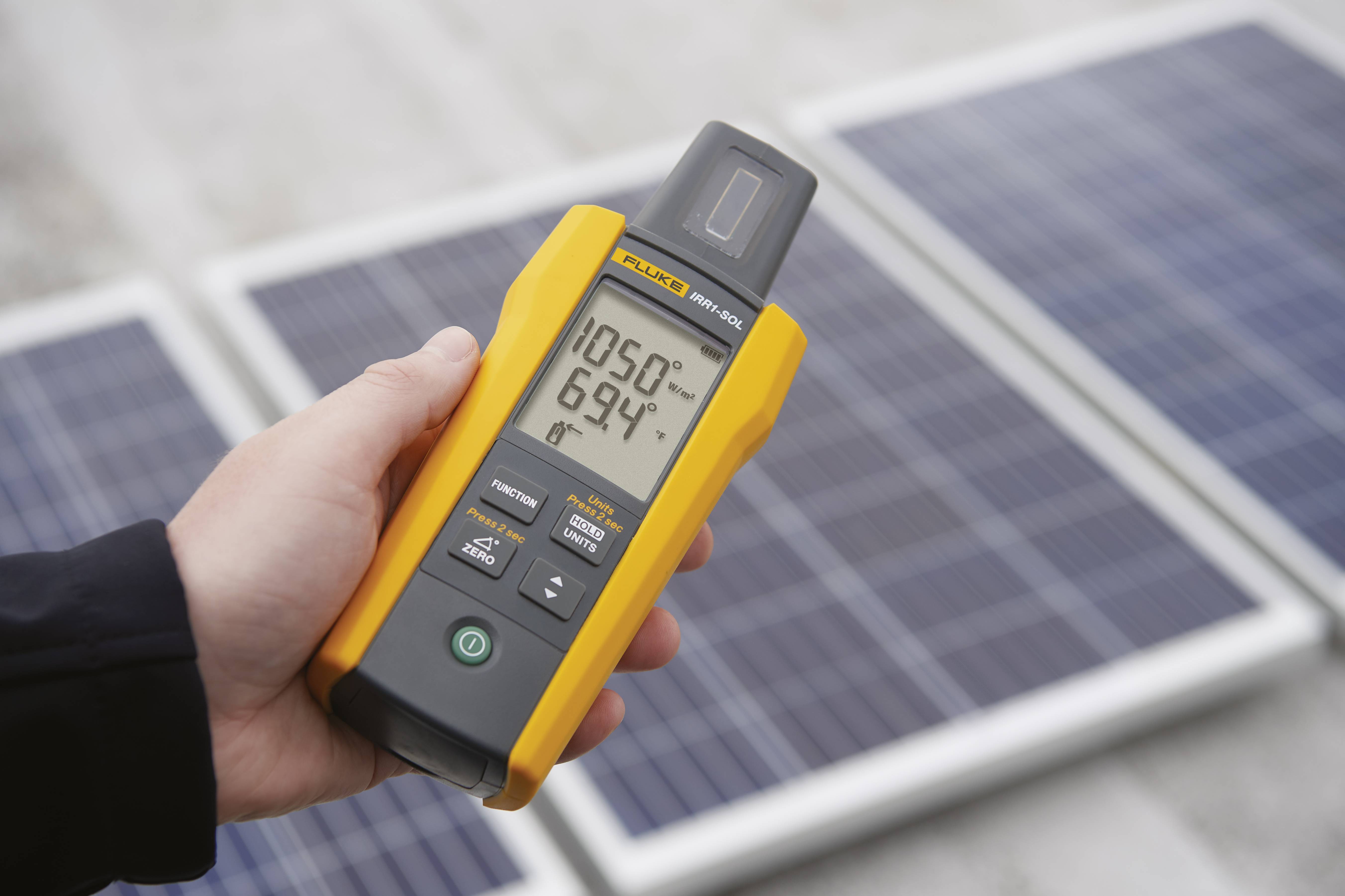 Een persoon houdt een draagbaar meetapparaat voor zonnepanelen. Het display toont meetwaarden voor zonnestraling en temperatuur.