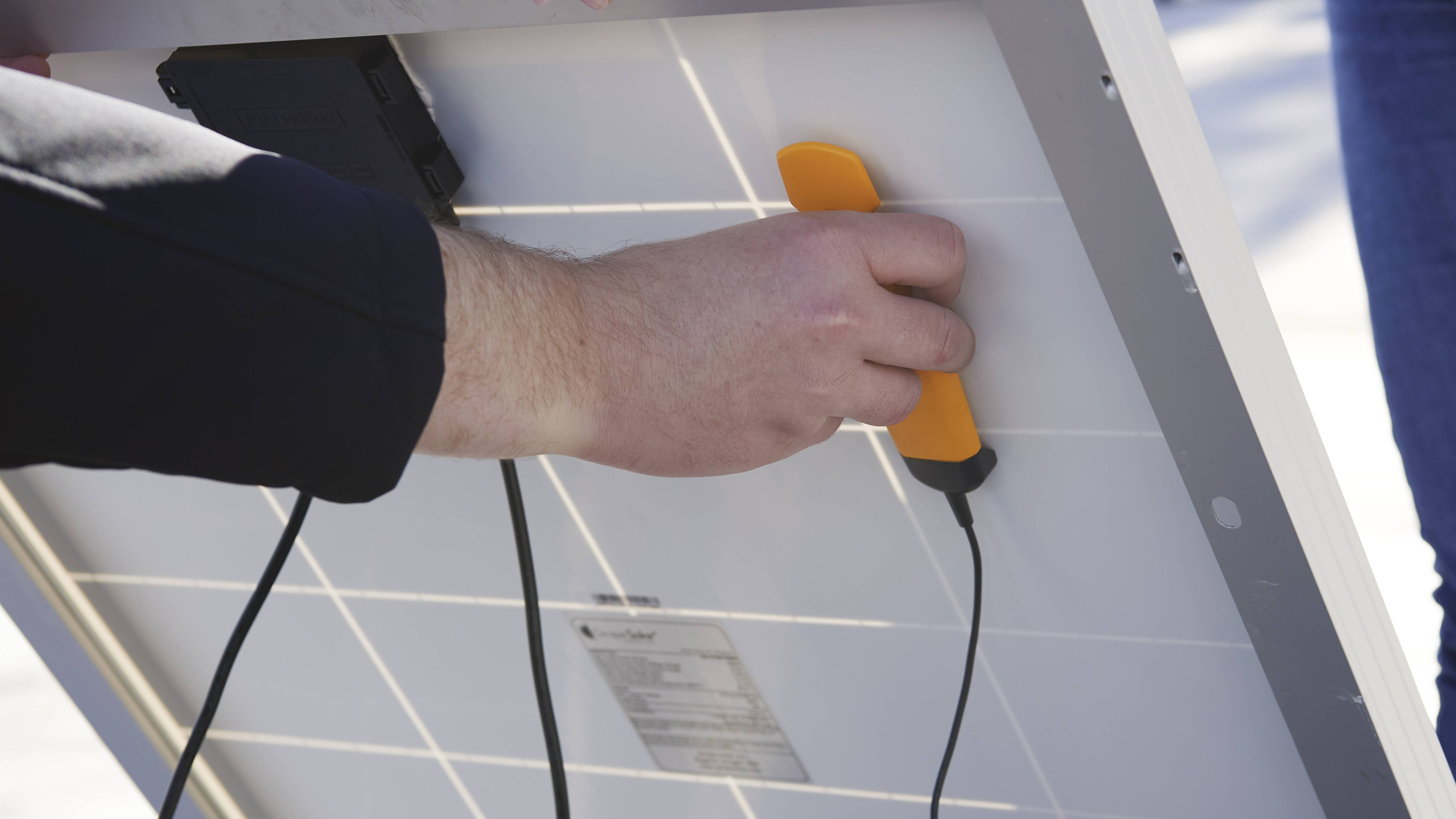 Een persoon houdt een zonnepaneel vast en gebruikt een oranje gereedschap. Kabels zijn aangesloten, wat wijst op een technische inspectie.