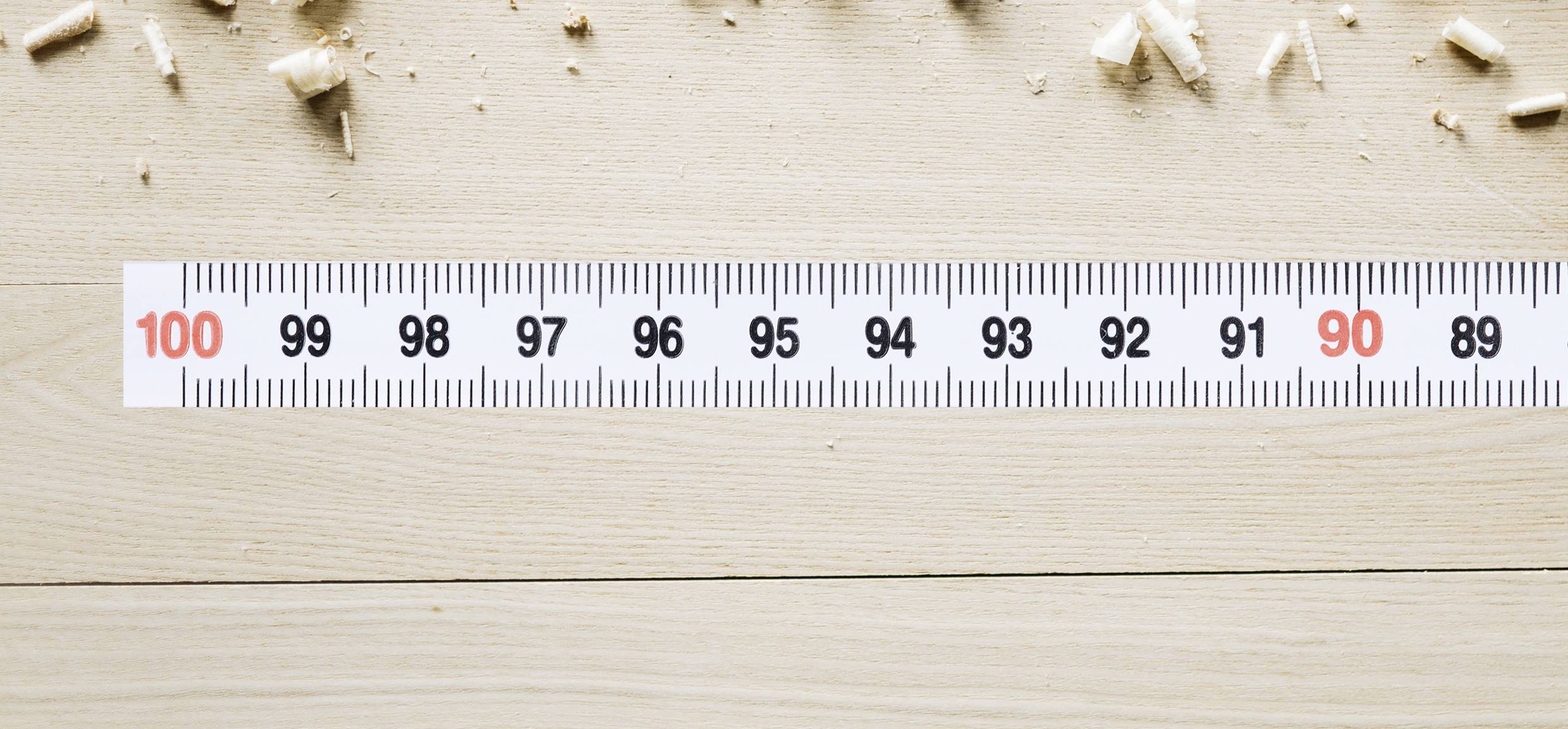 Een meetlint ligt plat op een houten vloer, met markeringen van 89 tot 100 zichtbaar. Houtspaanders zijn verspreid rond het meetlint.