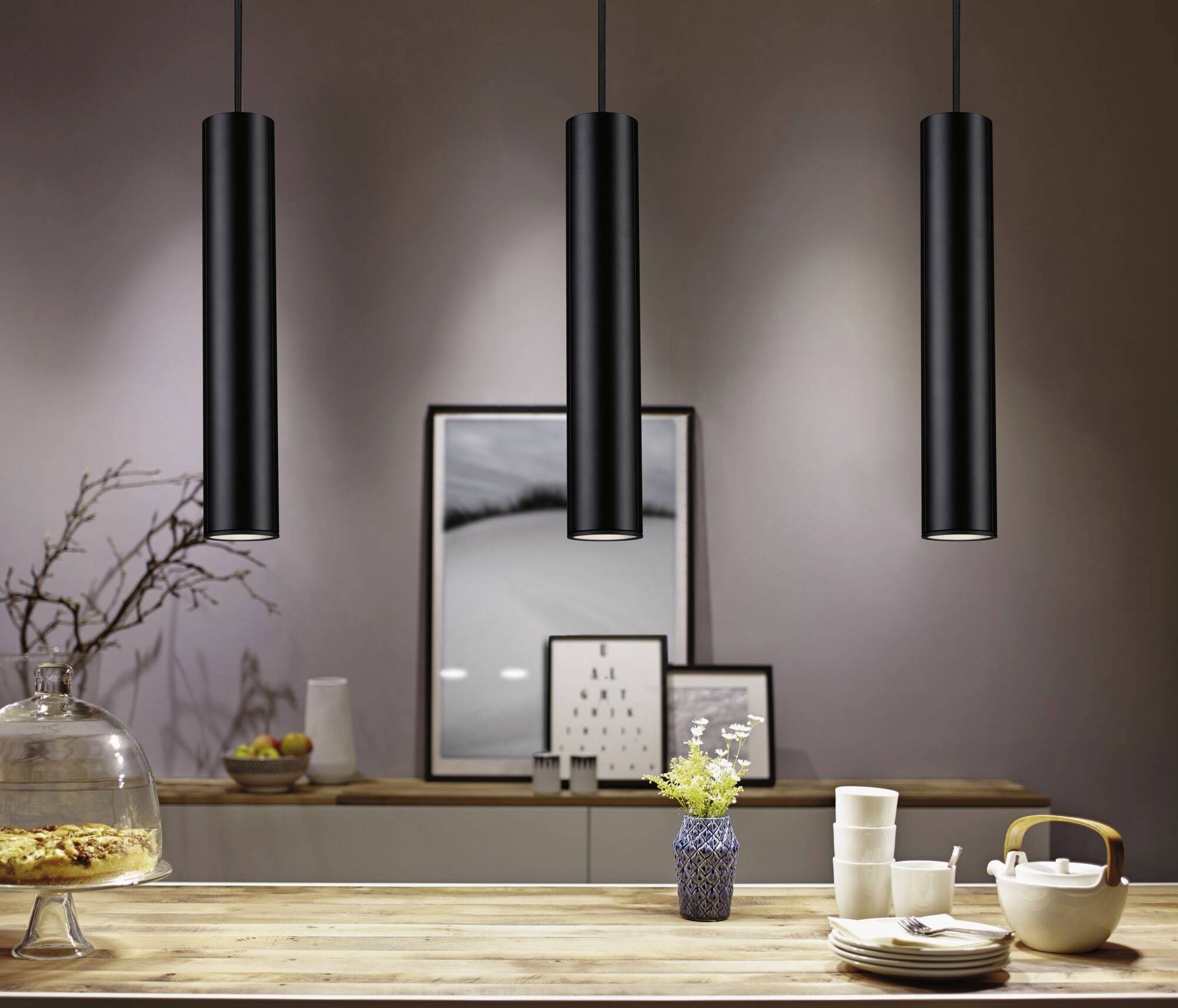 Een tafel met een bloemboeket, servies en een theepot, verlicht door drie moderne zwarte hangende lampen. Kunstwerk op de achtergrond.