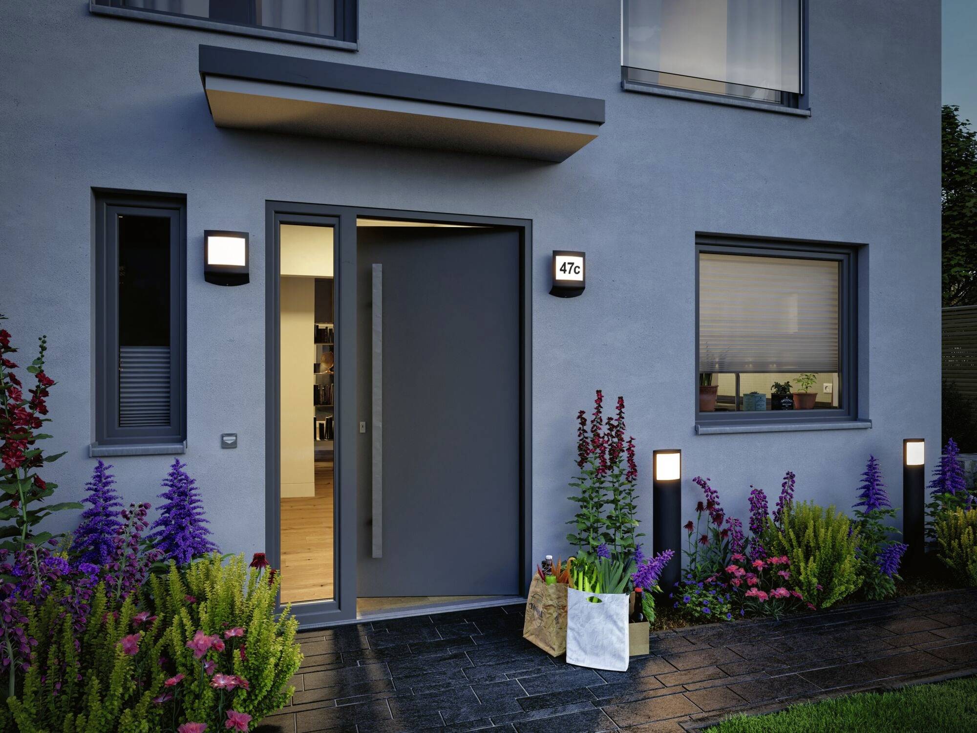 Een modern huis met een grijze gevel. De voordeur staat open, links en rechts bloeien planten. Op de traptrede staat een papieren tas.