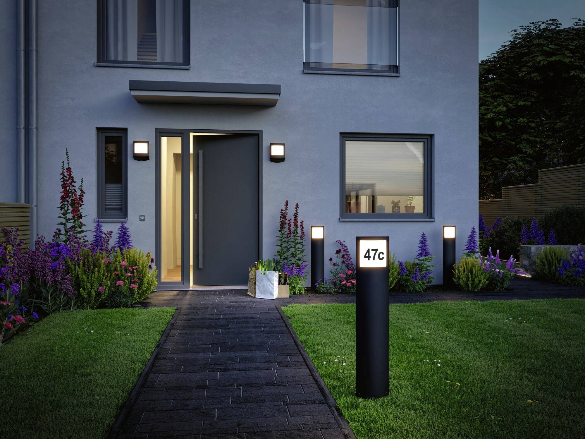 Een modern woonhuis met een openstaande deur, daarachter een verlicht pad en een tuin met bloemen. Een bord toont '47c'.