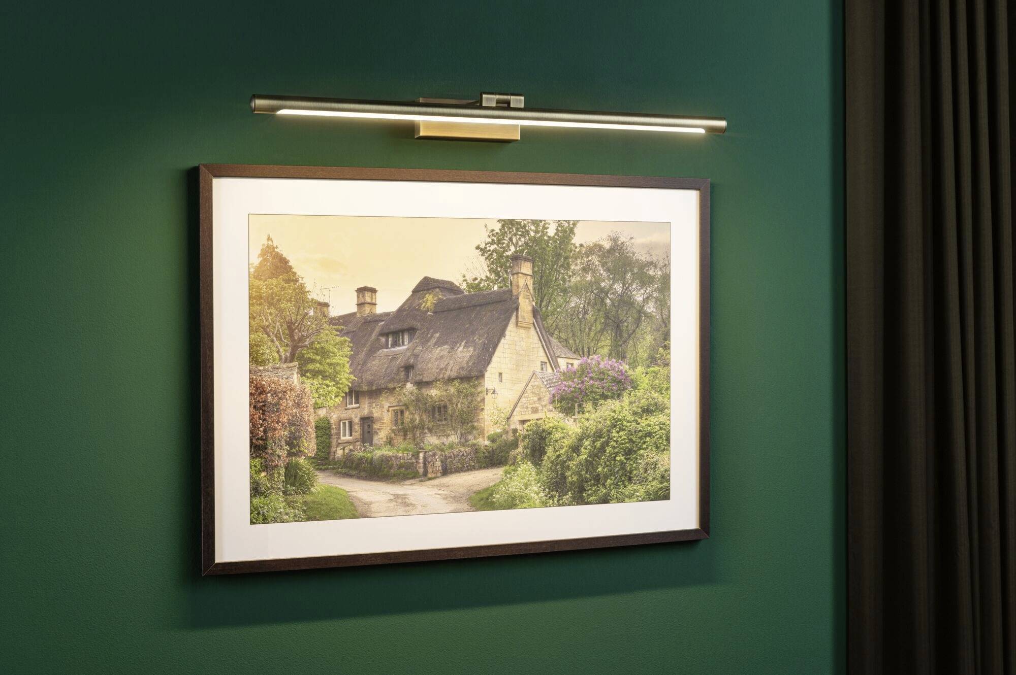 Een ingelijste foto van een schilderachtig, met stro gedekt huis, omgeven door bomen en struiken, hangt aan een groene muur.