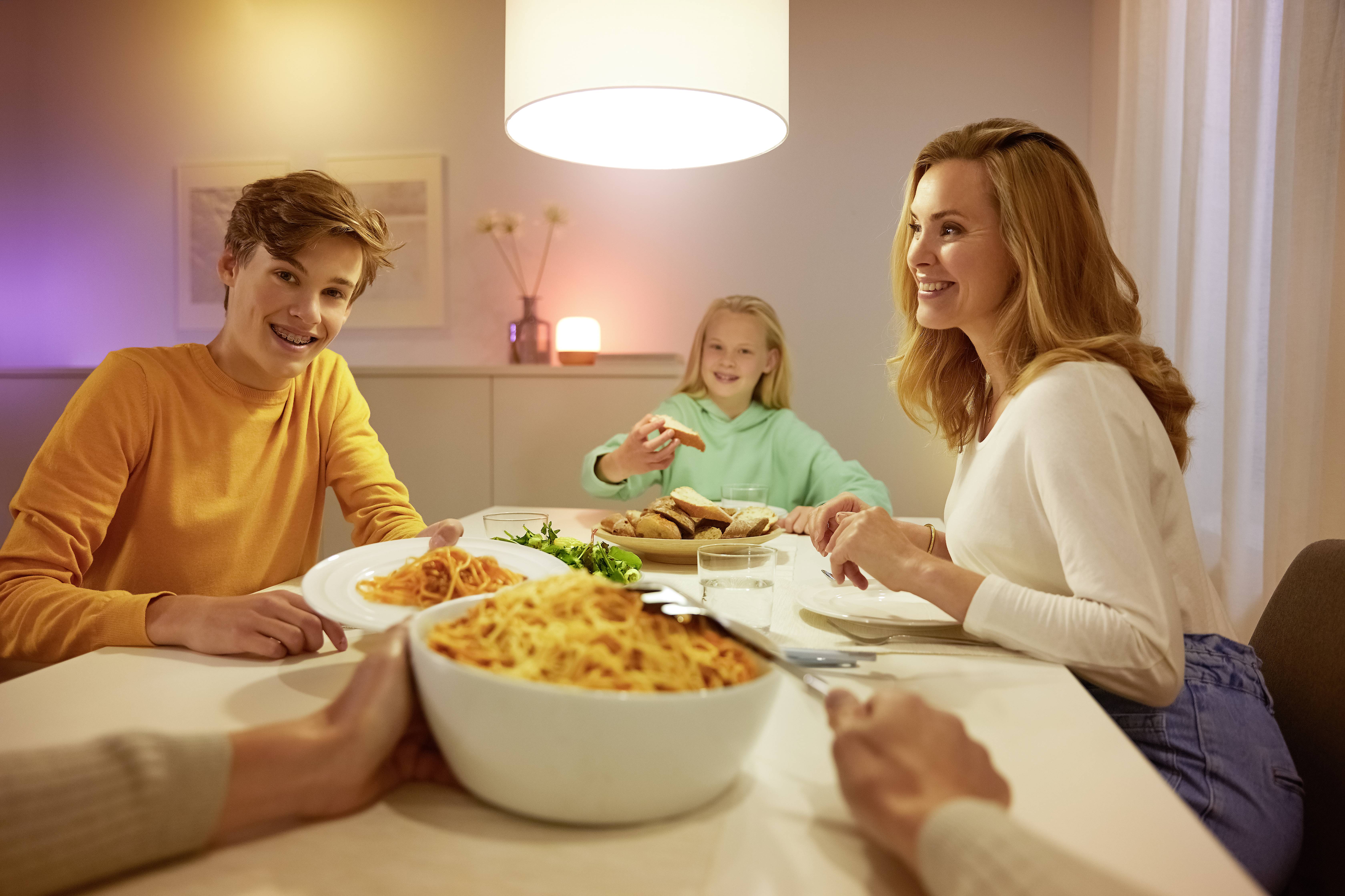 Een familie zit lachend aan tafel tijdens het avondeten. Een kom met pasta en borden met spaghetti staan voor hen. Ontspannen sfeer.